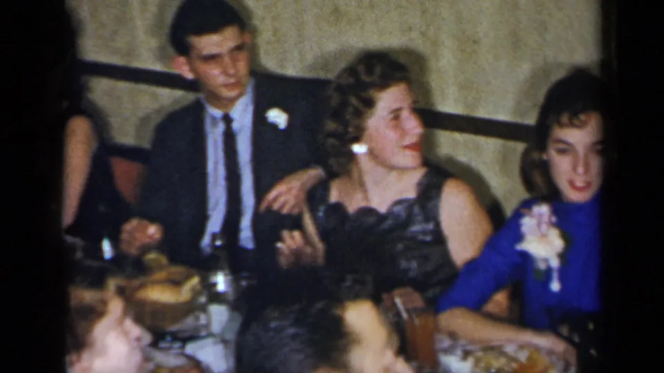 1954 New York home-movie frame showing young adults seated at a formal dinner table, with corsages, suits, food, and glasses visible.