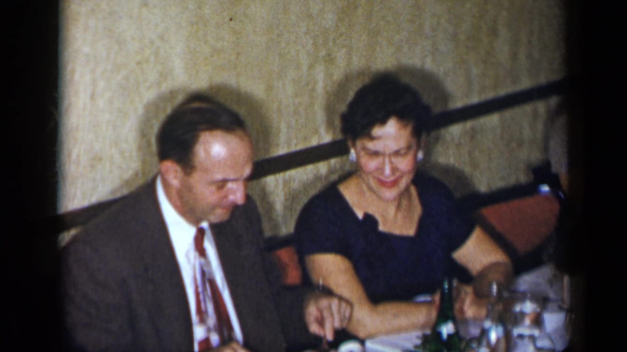 In 1954 New York home-movie footage, two adults sit at a table with drinks and glassware in a private interior.