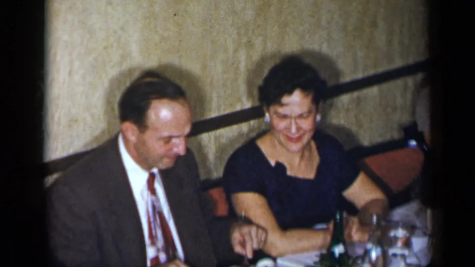 In 1954 New York home-movie footage, two adults sit at a table with drinks and glassware in a private interior.