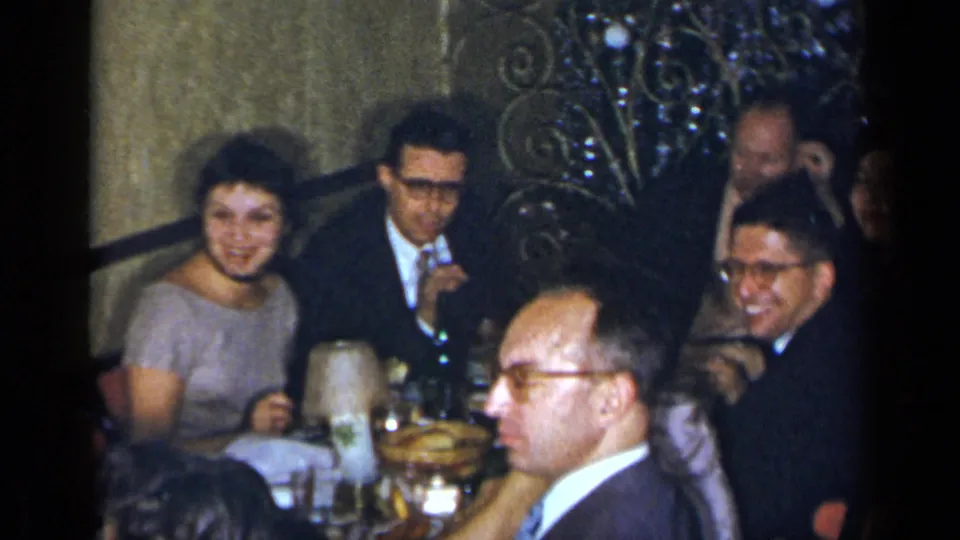 Adults in suits and dresses gather around a dining table with bottles, glassware, and food in a private New York interior.