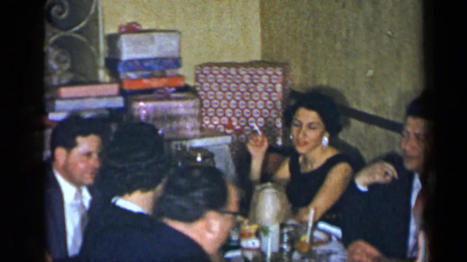 Guests sit around a table with wrapped gifts visible behind them, suggesting a celebration in the 1954 New York home movie.