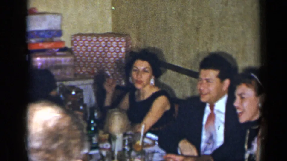 1954 New York home-movie frame of adults in formal clothing seated at a party table, with drinks and decor visible.