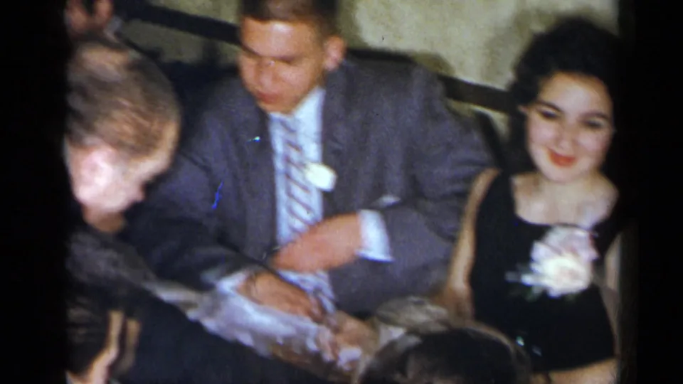 1954 New York home-movie frame showing a suited man, a woman in a dark dress with a white corsage, and nearby party guests.