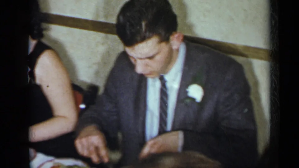 1954 New York indoor party frame with a suited man wearing a white boutonniere seated beside a woman in a dark dress.