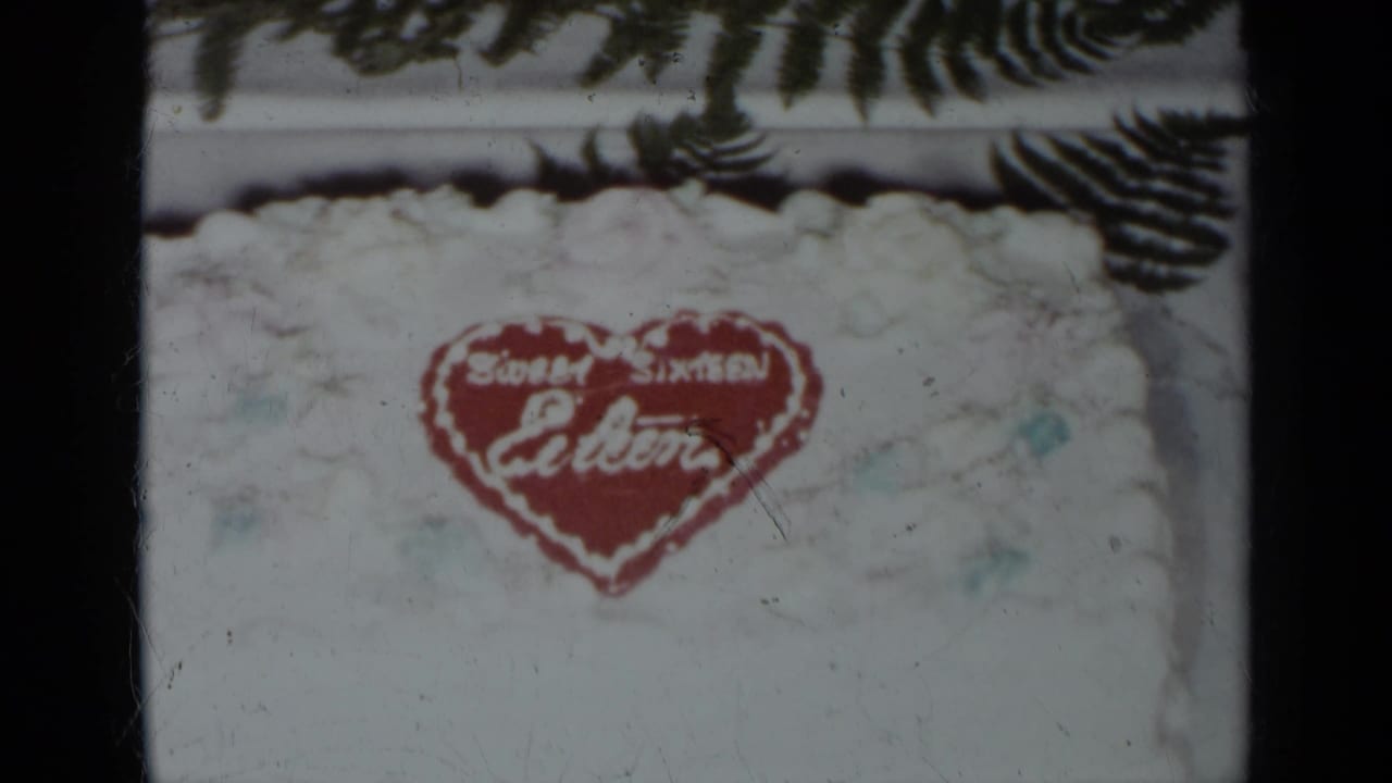 White frosted cake with a red heart reading Sweet Sixteen Eileen, framed by fern leaves.