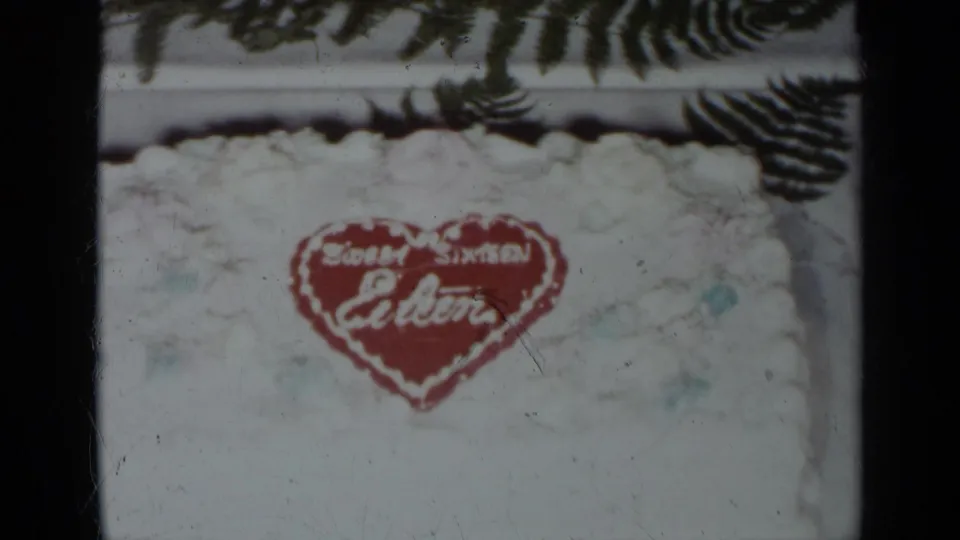 1954 New York 8mm frame showing a white frosted Sweet Sixteen cake with a red heart inscription and dark green fern leaves.