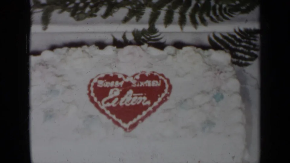1954 New York home-movie still of a white cake bearing a red Sweet Sixteen Eileen heart, bordered by fern leaves.