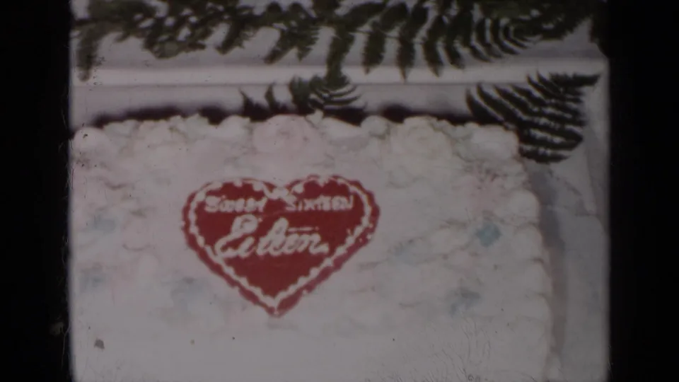 1954 New York 8mm frame of a white Sweet Sixteen cake with red heart lettering and fern leaves surrounding it.