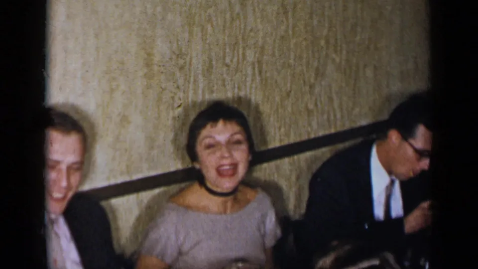 1954 New York home-movie frame showing adults in formal clothing socializing at a dining table in a dim restaurant interior.