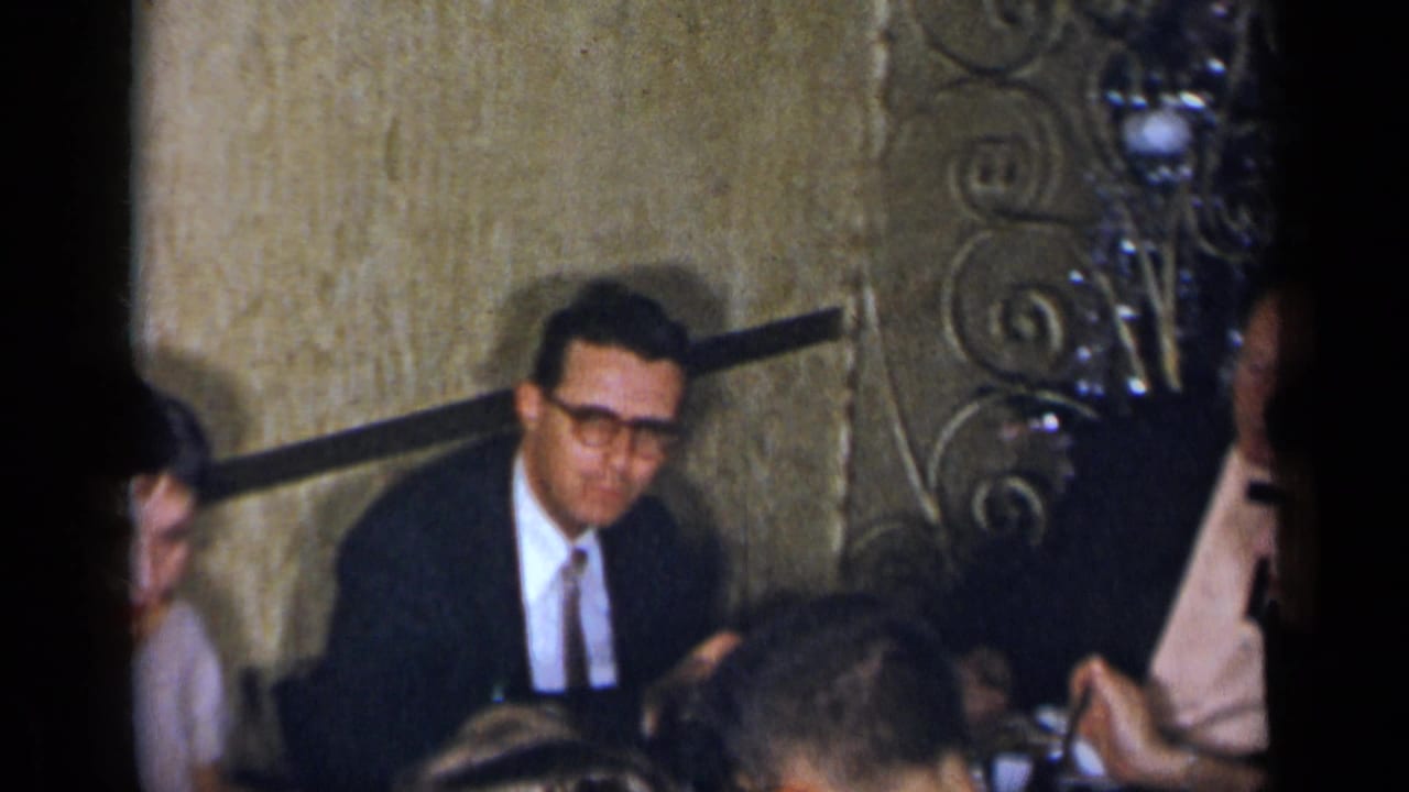 Men in suits and seated guests are seen beside a wall and ornate metalwork in a restaurant interior.
