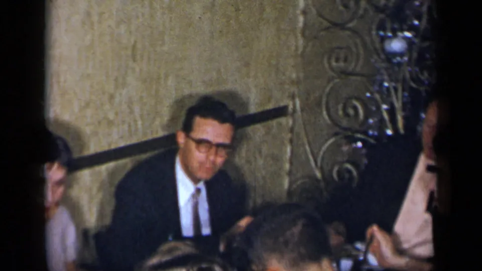 A 1954 New York restaurant gathering shows suited men and nearby guests seated around a dining table, partly obscured by the film frame edge.