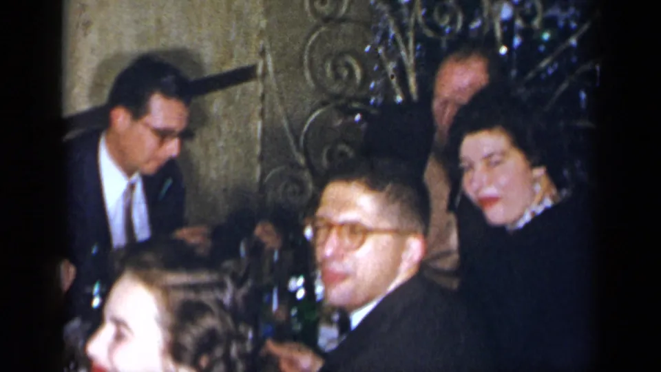 1954 New York home-movie view of adults at a crowded restaurant table with glassware, bottles, and formal mid-century clothing visible.