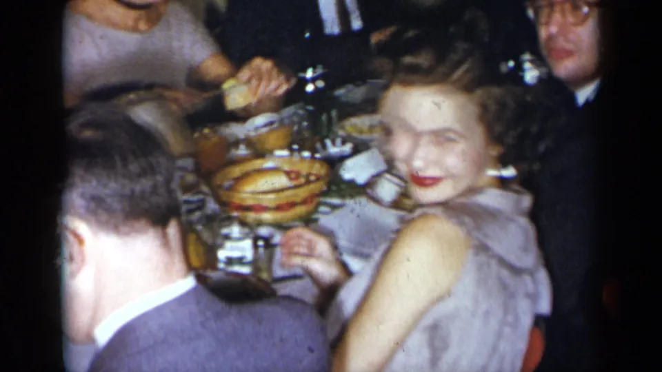 A woman in a light dress smiles toward the camera at a 1954 New York restaurant table crowded with food, drinks, and seated guests.
