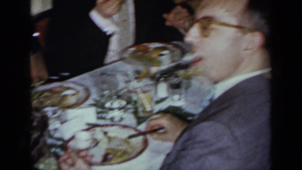 Man in suit and glasses turns toward the camera while seated at dinner.