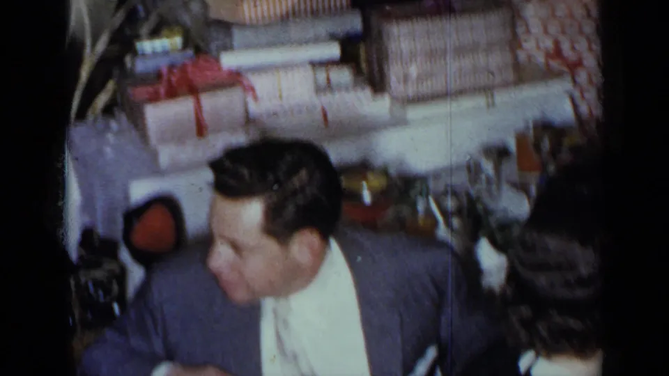 1954 New York home-movie frame with a seated man near a dining table and wrapped gifts stacked behind him.