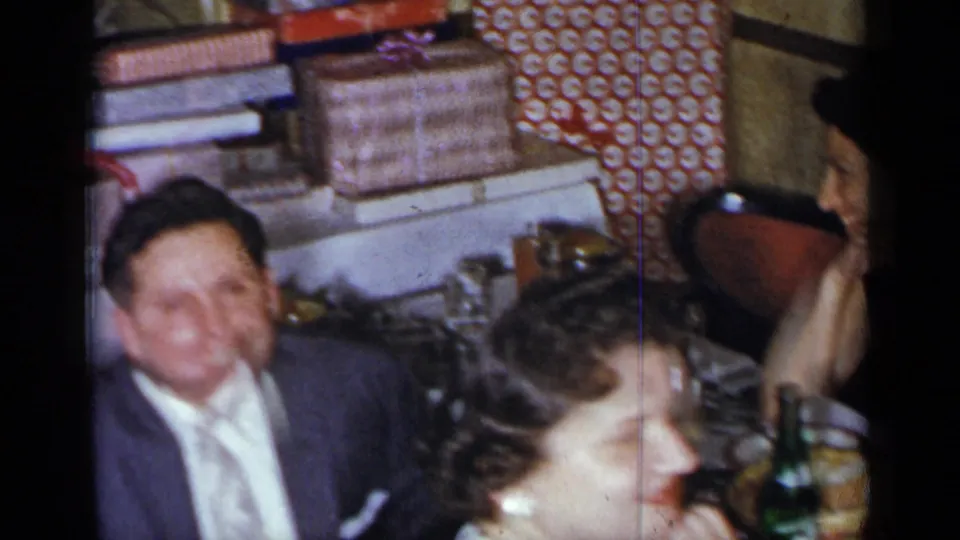 1954 New York home-movie frame showing adults smiling and talking near a table with wrapped gifts behind them.