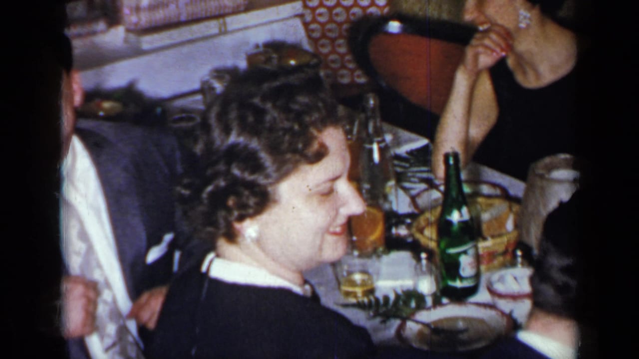 Guests seated at a dinner table with plates, glasses, bottles, and a smiling woman in dark clothing.