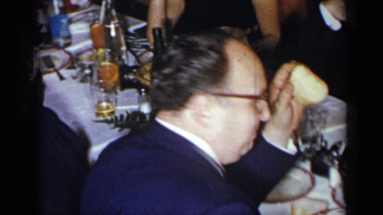 Man in a suit seated beside a dinner table with plates, glasses, bottles, and other guests nearby.