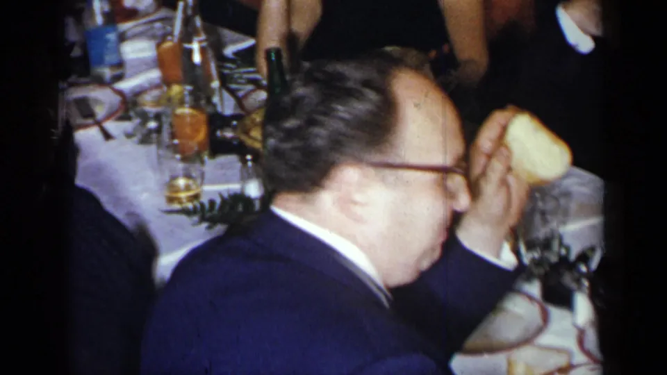 1954 New York dinner-party frame with a suited man in profile, table settings, drinks, and partially visible guests.