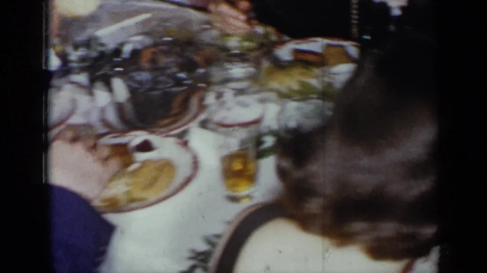 Handheld 8mm frame from a 1954 New York dinner showing table settings, bread rolls, drinks, and guests in motion blur.