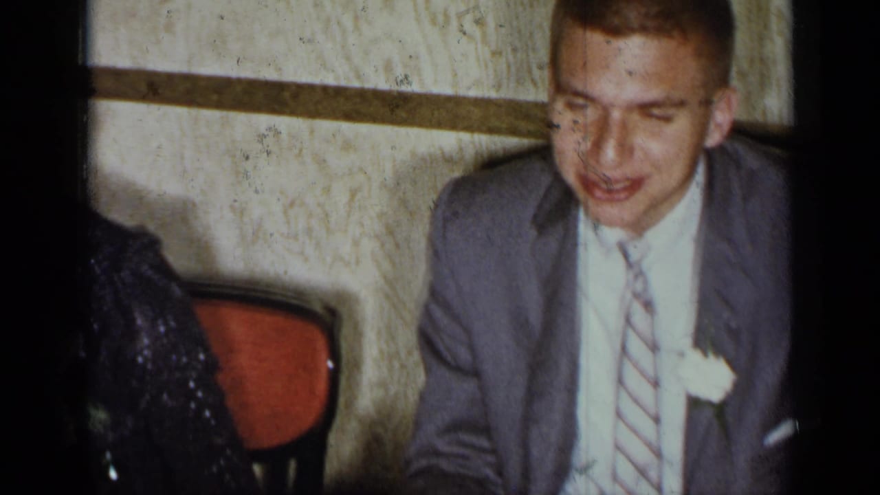 Young man in a suit sits at a dinner table beside partially visible guests.