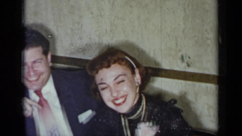 The couple laughs together at the table, with the woman's pearl headband and corsage visible in the 1954 home-movie frame.