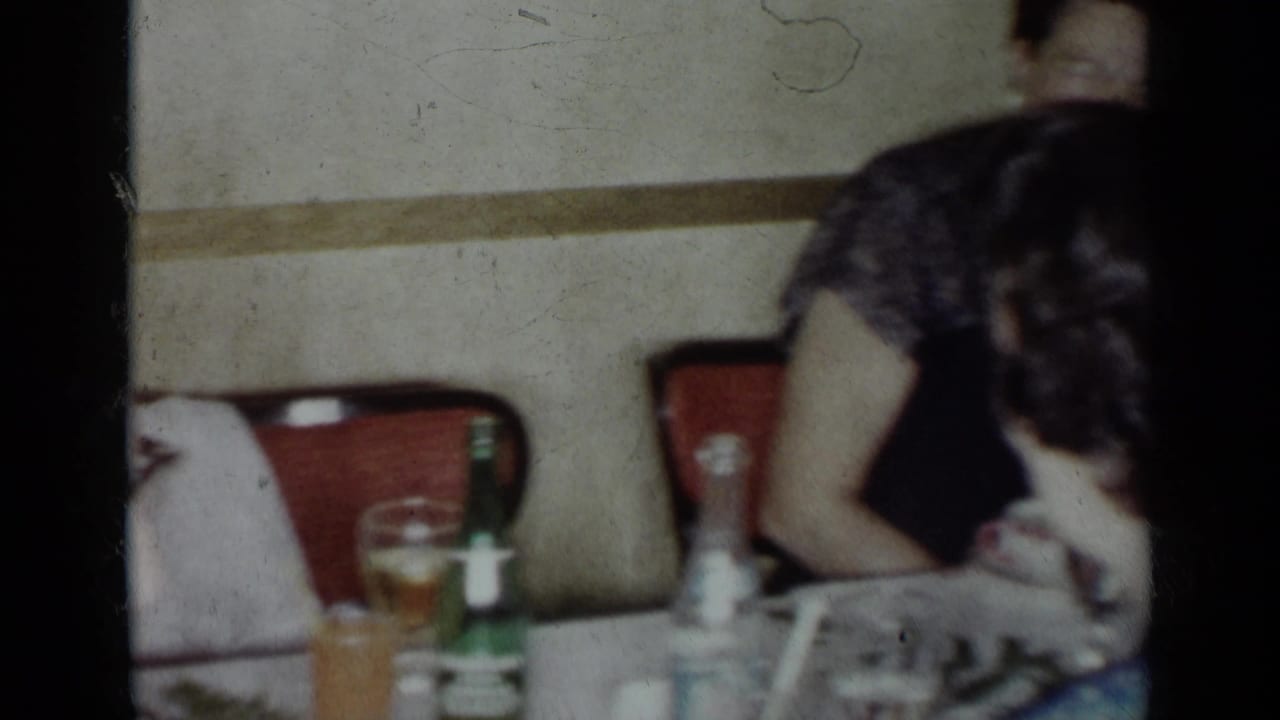 1954 New York home-movie frame showing adults in dress attire seated at a dining table with bottles, glasses, and red chairs.