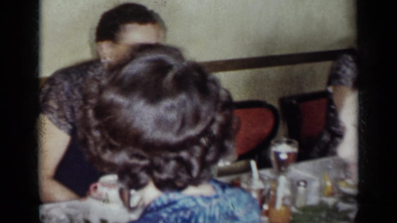 A 1954 New York dinner scene with a woman in blue seen from behind, other adults, red chairs, glasses, and bottles on the table.