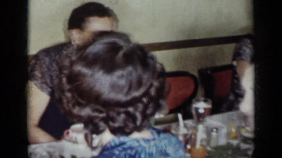 A 1954 New York dinner scene with a woman in blue seen from behind, other adults, red chairs, glasses, and bottles on the table.