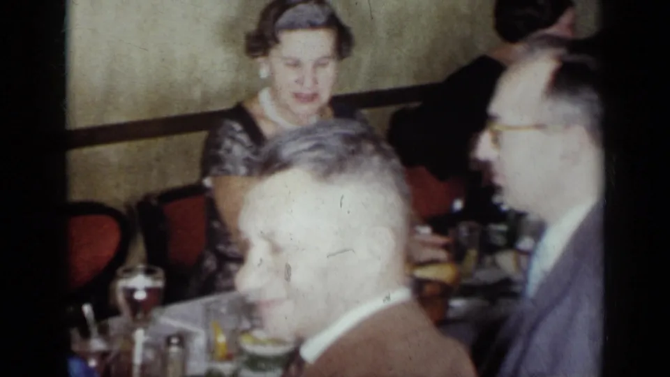 1954 New York home-movie still of adults at a crowded dining table, with a woman in a patterned dress and men in suits nearby.