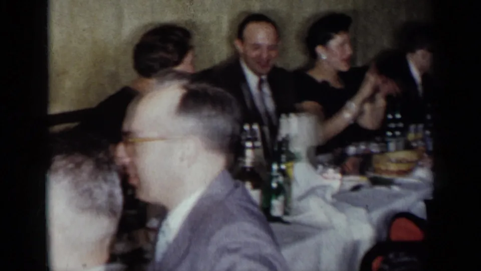 A 1954 New York dinner gathering shows several adults in formal attire seated along a long table with bottles, glasses, and plates.