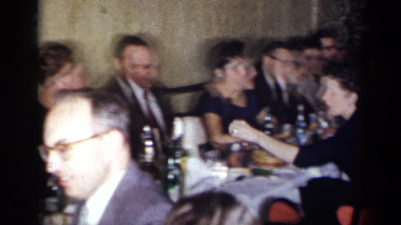 1954 New York home-movie frame of adults in suits and dresses seated around a restaurant dinner table with bottles, glasses, and plates.