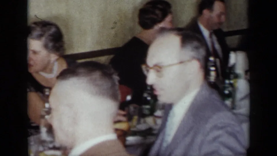 1954 New York restaurant dinner scene with adults seated closely, tableware and bottles visible, and several faces turned in conversation.