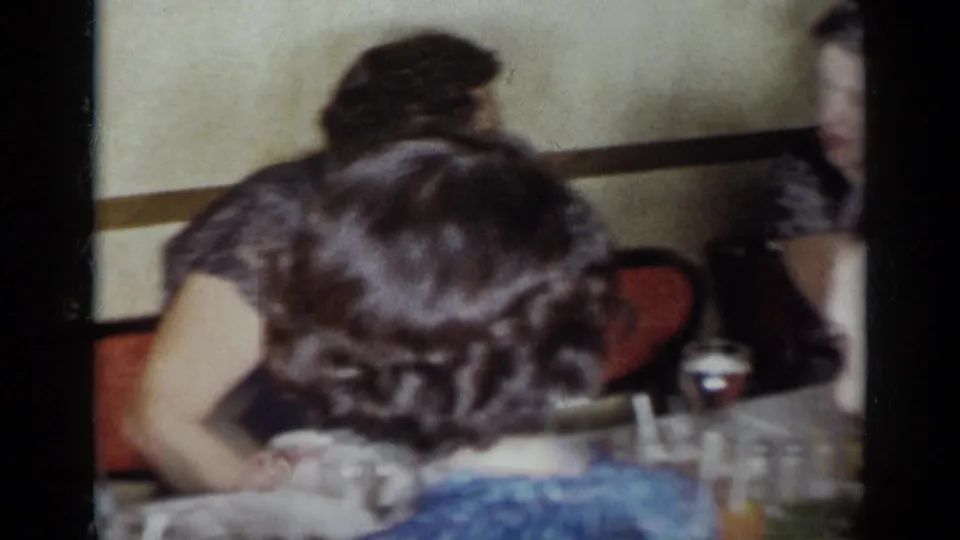 Close 8mm view of women at a 1954 New York restaurant table, with glassware, plates, red chairs, and a softly lit interior wall.