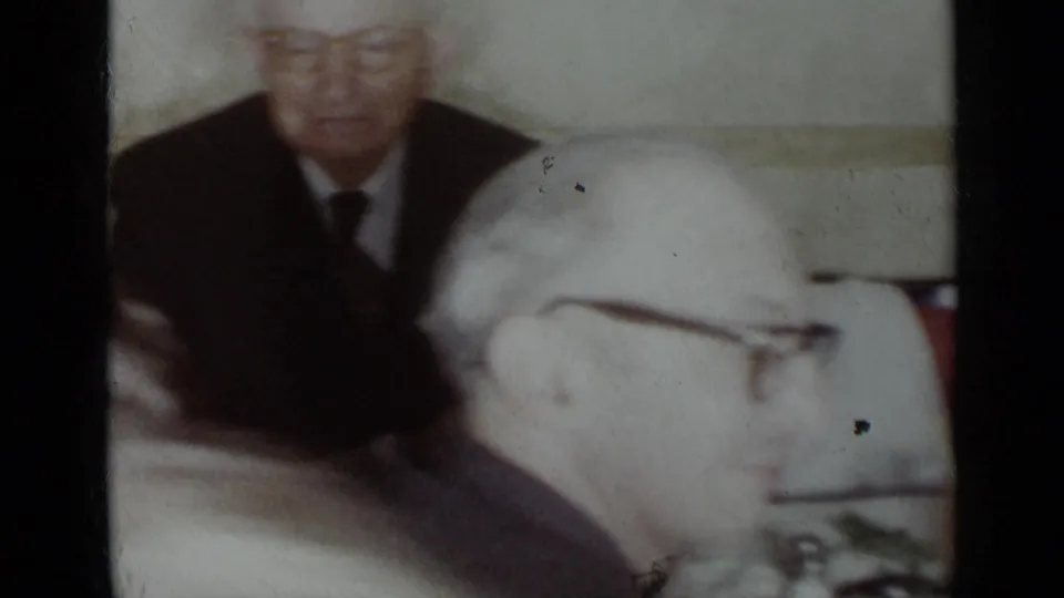 Soft 1954 New York home-movie frame of older men at a restaurant dinner table, with eyeglasses, suits, and blurred tableware visible.