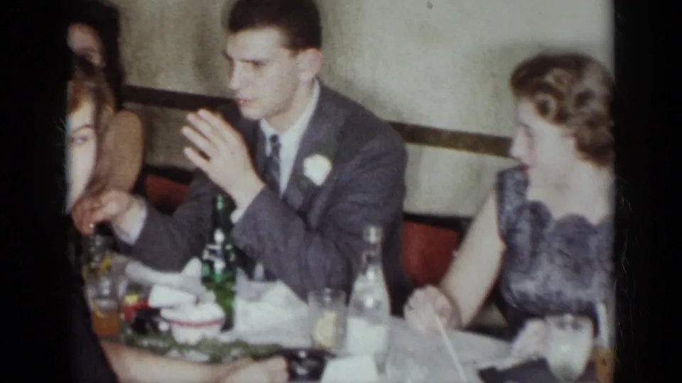 A suited man with a boutonniere sits beside women at a formal New York restaurant dinner, with bottles, glasses, and napkins on the table.
