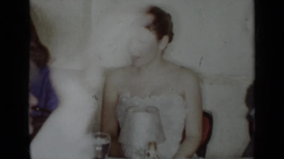 In 1954 New York home-movie footage, a woman in a white dress sits at a formal table with partially visible guests nearby.
