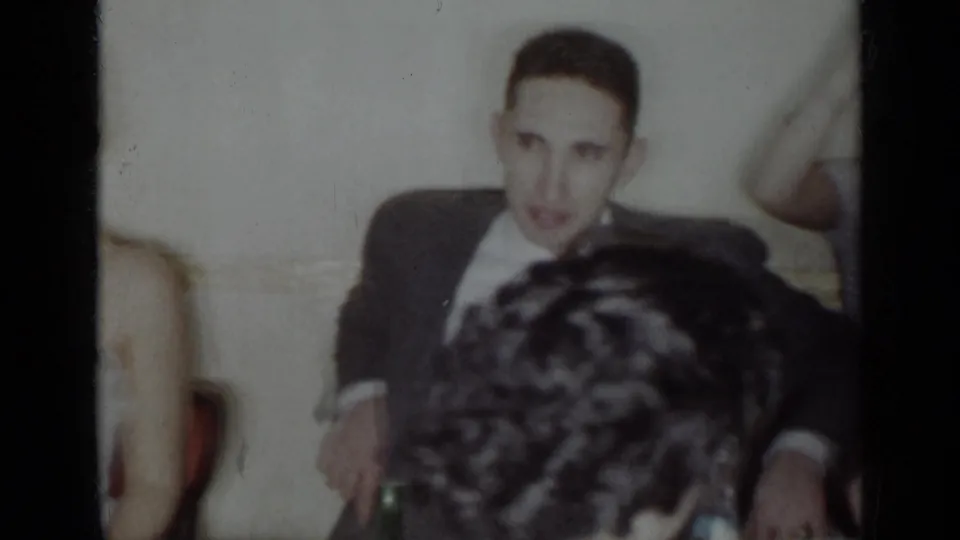 A man in a dark suit faces the camera during a formal 1954 New York indoor gathering, with other guests partly blocking the view.