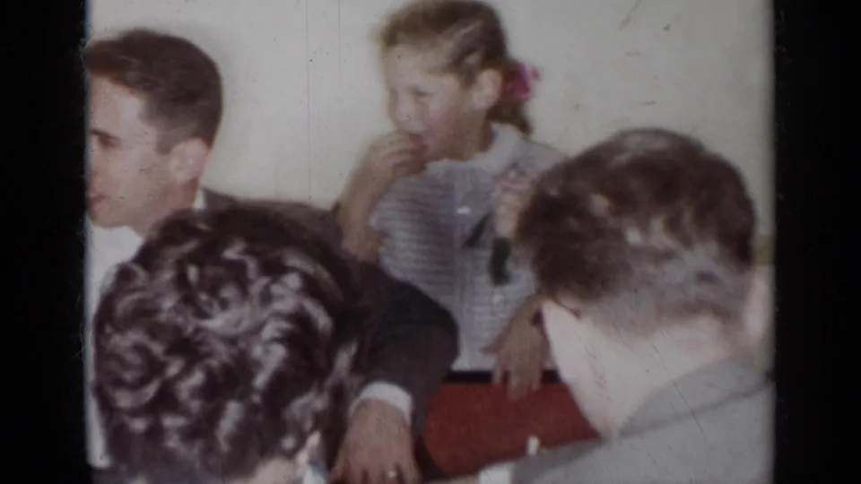 Guests in formal clothing sit at a 1954 New York gathering as a young girl with a hair ribbon eats near the table.