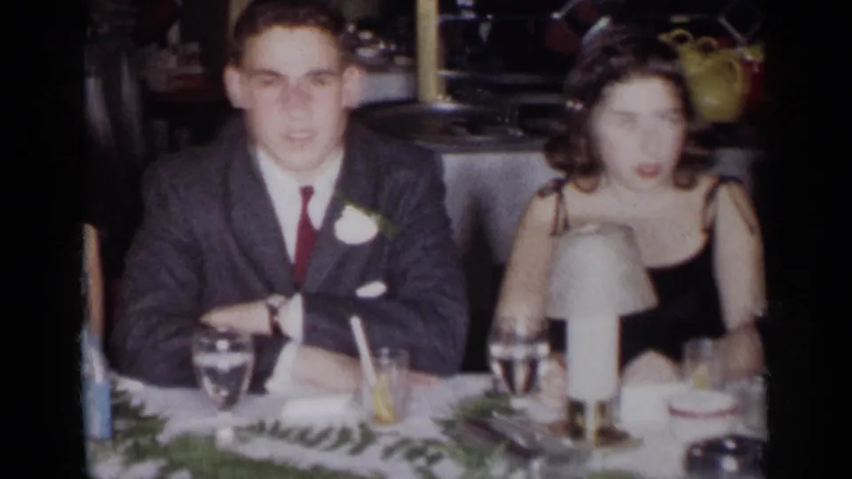 1954 New York home-movie frame showing a young man with a boutonniere and a woman in a black dress at a decorated table.