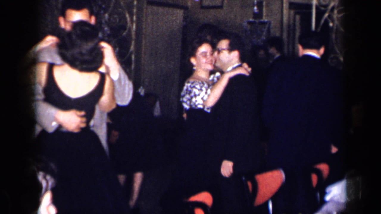 1954 New York home-movie frame showing adults dancing closely in a dim interior with suits, dresses, and red chairs visible.