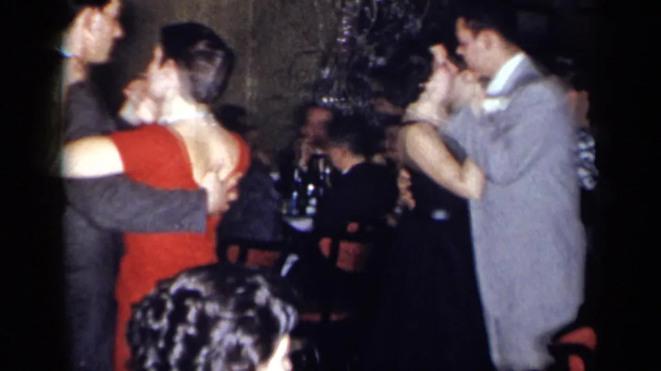 A woman in a red dress dances with a partner in a dim 1954 New York party interior, with seated and standing guests around them.