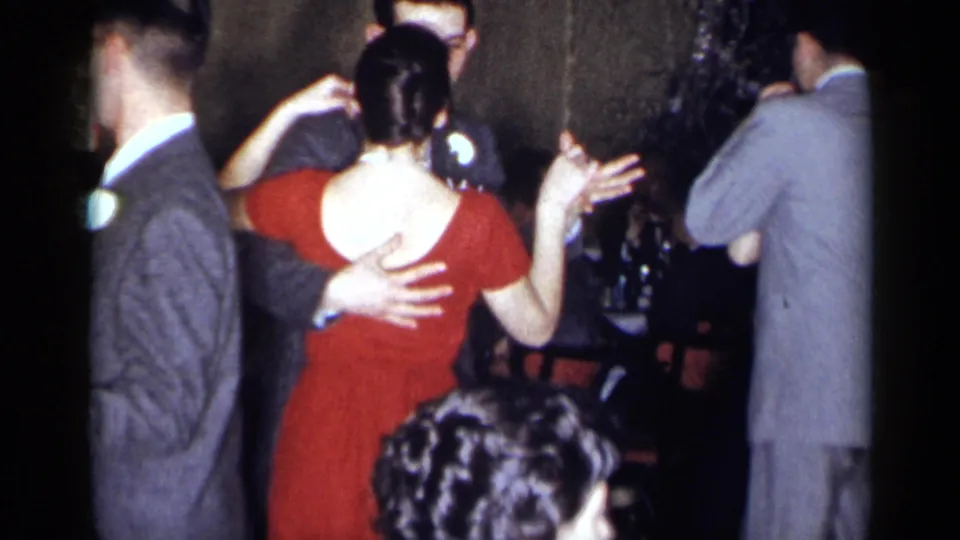 Party guests dance in pairs in a 1954 New York home-movie frame, with a red dress, gray suit, and crowded interior visible.