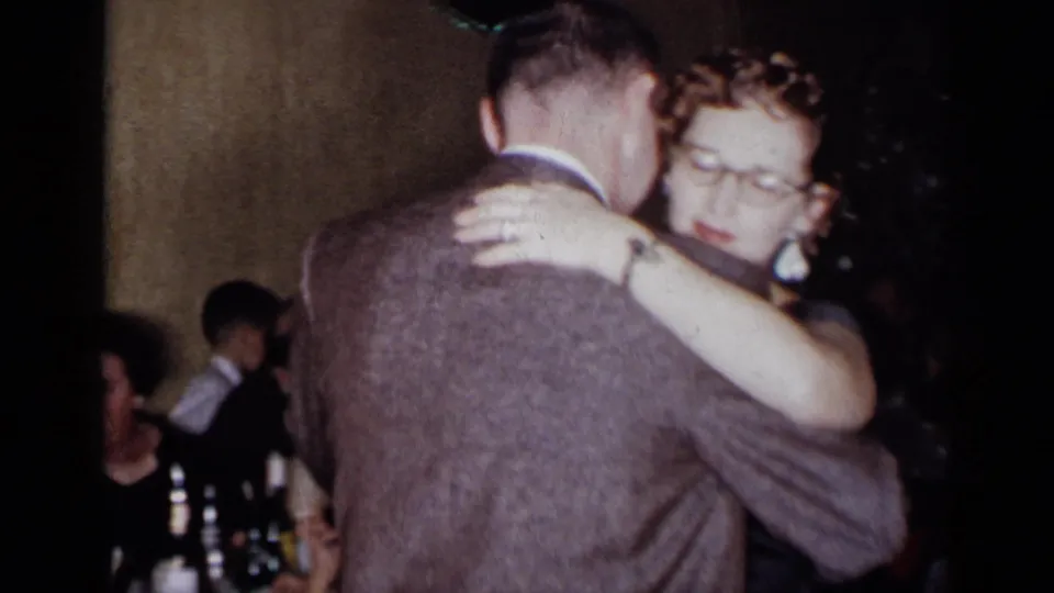 The woman, wearing glasses and a dark dress, dances closely with a suited man as seated guests remain visible behind them.