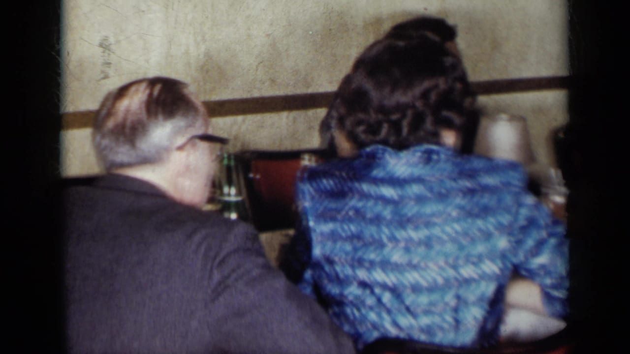 1954 New York home-movie frame showing adults seated at a formal table with glassware and a woman in a blue dress facing away.