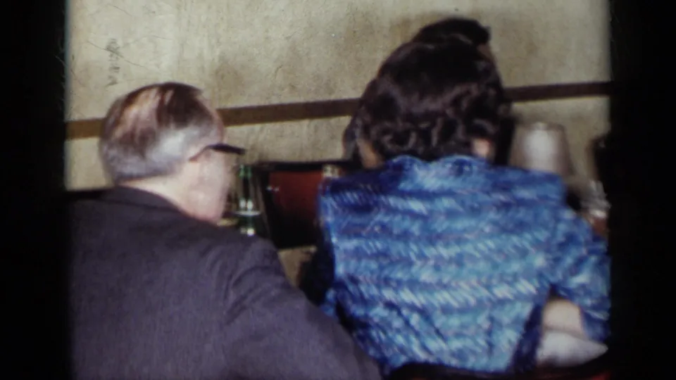 1954 New York home-movie frame showing adults seated at a formal table with glassware and a woman in a blue dress facing away.