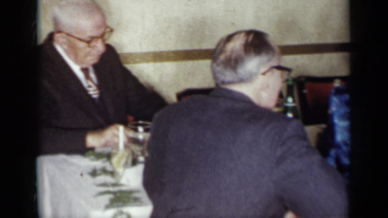 Two older men in suits sit at a table with glasses and a white tablecloth.