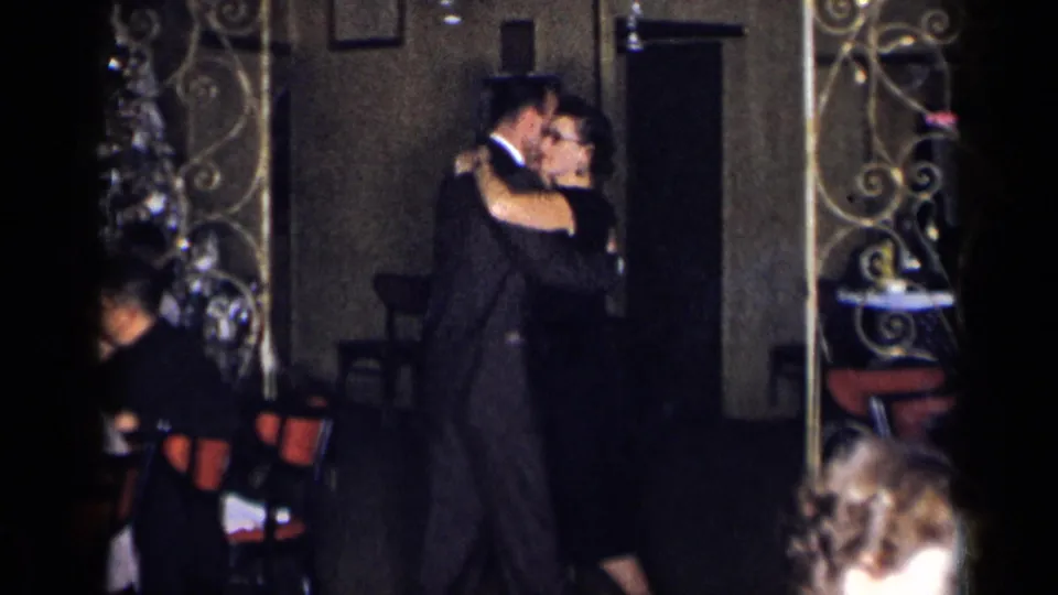 1954 New York home-movie frame showing a dark-suited man and woman dancing beside ornate room dividers and chairs.