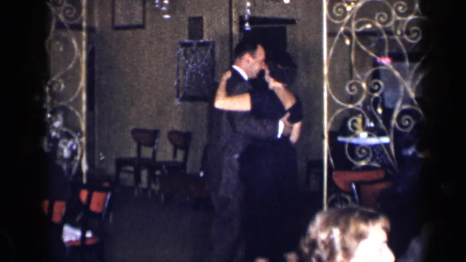 1954 New York home-movie frame of a couple dancing closely in a dim formal room with decorative partitions visible.
