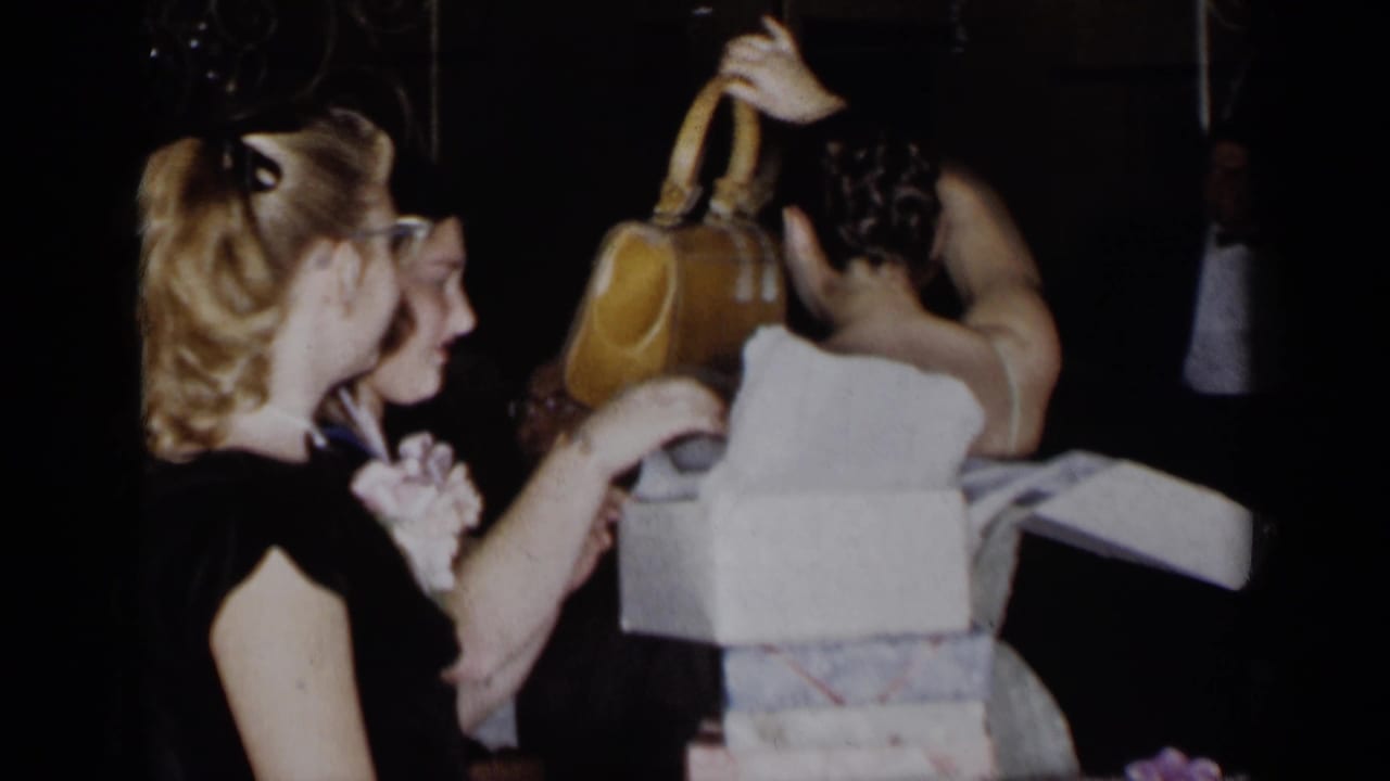Woman in black dress watches another woman open a white gift box at a dim formal party.
